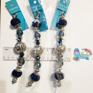 Può includere: Tre perline da pranzo di "hildie & jo" in confezioni di plastica trasparente. Le perline sono blu navy, argentate e trasparenti. Le perline sono disposte verticalmente. Un righello è posto accanto alle perline.
