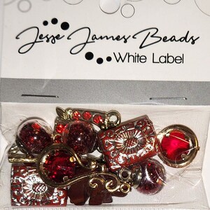 Può includere: Un sacchetto di plastica trasparente contiene una collezione di perline rosse e dorate. Le perline includono forme rotonde, quadrate e decorative. La borsa ha il testo "Jesse James Beads White Label" stampato.