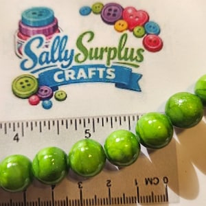 May include: Close-up of a strand of green beads with a marbled effect. A ruler shows the beads are approximately 0.75 inches in diameter. The image also shows the text "Sally Surplus Crafts" with decorative buttons.