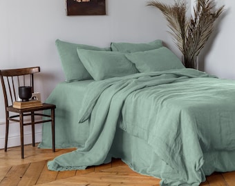 May include: A bed dressed with sage green linen bedding, including a duvet cover, pillows, and a bed skirt. The room features a wooden floor, a wooden chair, and a large mirror. A painting hangs on the wall.