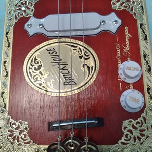 May include: A red wooden cigar box guitar with gold accents and a white pickup. The guitar has a volume and tone knob. The cigar box has the text "Brick House" and "Juman Cigar Co. - Nicaragua" on it.