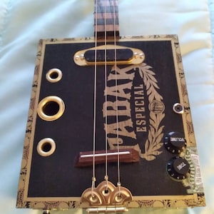 May include: A cigar box guitar with a black body and gold accents. The guitar has a wooden neck and a gold-colored bridge. The body of the guitar is decorated with a gold-colored label that says "PALMAR ESPECIAL". The guitar has two knobs on the side, one for volume and one for tone.