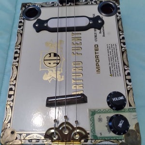 May include: A white cigar box guitar with gold accents and black knobs. The guitar has a black pickguard and a label that reads "ARTURO FUENT IMPORTED CURLY HONDURAS DOMINICAN REPUBLIC".