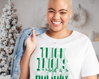 Puede incluir: Camiseta blanca con el texto "THICK THIGHS and HOLIDAY VIBES" en verde. La camiseta tiene cuello redondo y mangas cortas. Una chaqueta vaquera azul claro está sobre el hombro.