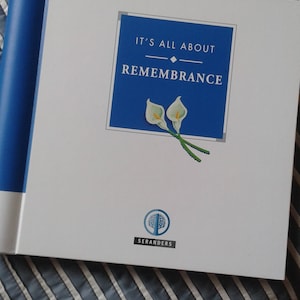 May include: A white book with a blue spine and a blue square on the front. The square has the text "It's All About Remembrance" and a picture of two white calla lilies. The book has the logo "Seranders" at the bottom.