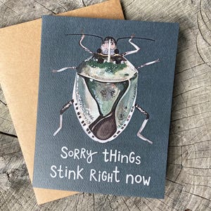 Könnte beinhalten: Eine dunkelgraue Grußkarte mit einer Aquarellillustration einer grünen und braunen Stinkwanze. Der in Weiß am unteren Rand der Karte gedruckte Text lautet, Sorry things STINK Right now. Ein schlichter brauner Umschlag liegt hinter der Karte.