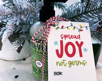 Puede incluir: Una botella verde de jabón de manos con una etiqueta blanca y un lazo de cuerda rojo y blanco. Una etiqueta de regalo blanca con el texto "Spread Joy not germs" y "From:" está unida a la botella.