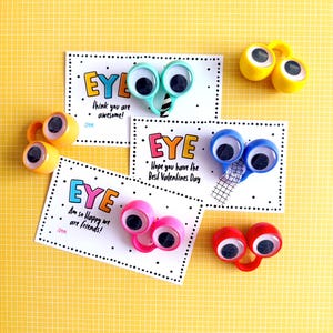 Può includere: Una collezione di accessori a forma di occhio e biglietti di auguri. Le carte presentano la parola "EYE" in lettere colorate, con messaggi come "think you are awesome!" e "Am so Happy we are friends!" Gli occhi sono di vari colori.