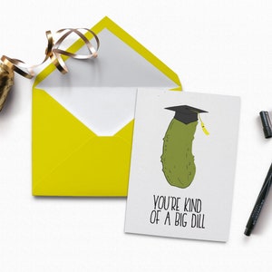 Peut inclure: Une carte de graduation avec un cornichon vert portant un bonnet de graduation noir et un gland jaune. La carte dit "You're kind of a big dill".