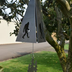 May include: A metal wind chime with a triangular shape. The chime features a silhouette of a Bigfoot and a tree design. The chime is hanging from a tree branch.