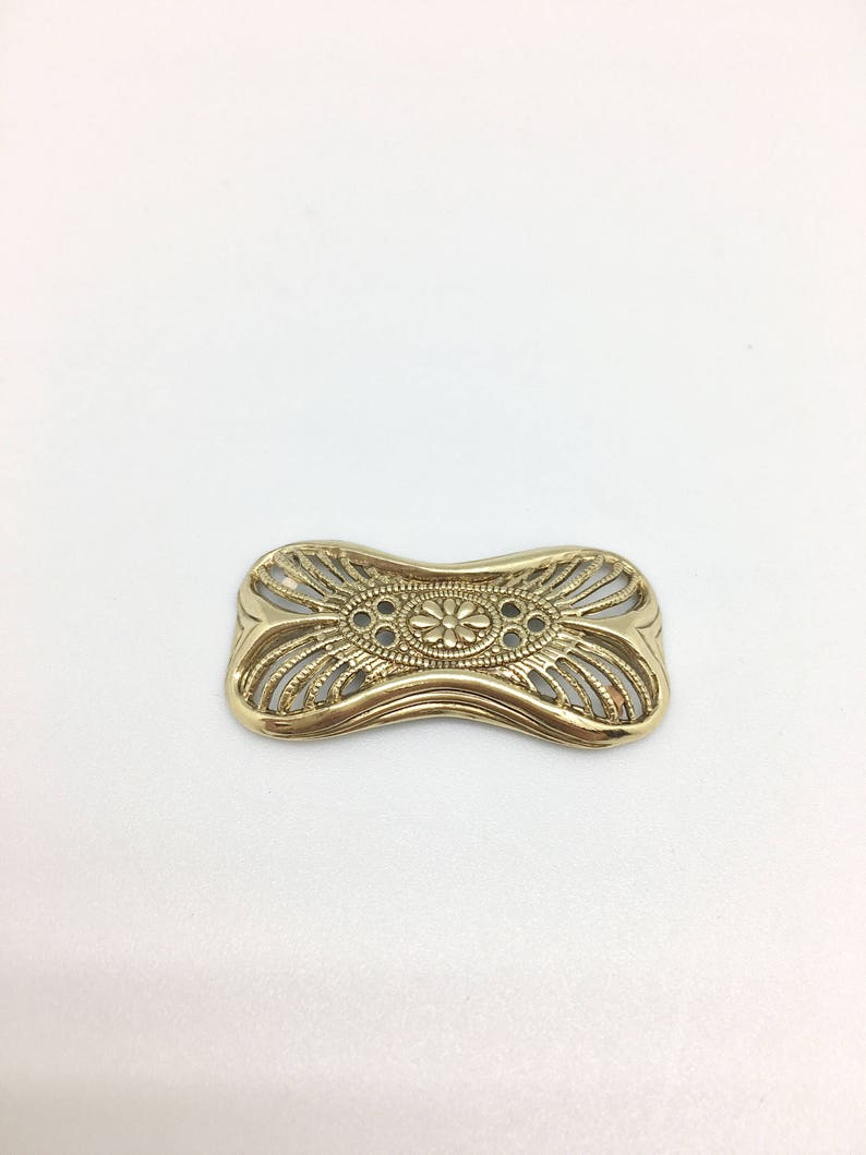 May include: A gold-toned metal belt buckle with a decorative, intricate design. The buckle has a rounded, slightly curved shape and features a central floral motif.