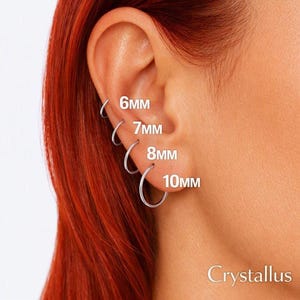 May include: Four silver-toned hoop earrings arranged in a vertical row on an earlobe. The hoops are labeled with their respective sizes: 6mm, 7mm, 8mm, and 10mm. The text Crystallus is visible in the bottom right corner of the image.