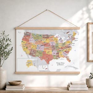 May include: A wall-hanging map of the United States of America printed on paper, secured between two light-colored wooden rails with a string for hanging. The map features colorful state borders, illustrations of regional landmarks, and text.