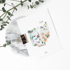 May include: A white rectangular art print featuring a watercolour map of Ohio. The map includes illustrations of trees, a deer, a squirrel, a sailboat, a blimp, and buildings. The word Ohio is printed in small script at the bottom centre of the paper.