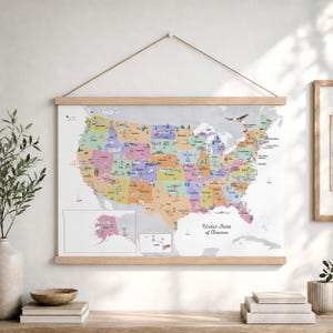 May include: A wall-hanging map of the United States of America featuring colorful state sections with illustrated landmarks, wildlife, and text labels. The map is secured between two light-toned wooden rails and suspended by a thin brown cord.