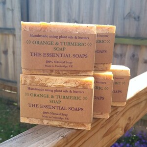 May include: Four bars of handmade orange and turmeric soap, stacked on a wooden surface. The soap is labelled "The Essential Soaps" and is made with plant oils and butters. The soap is 100% natural and made in Cambridge, UK.