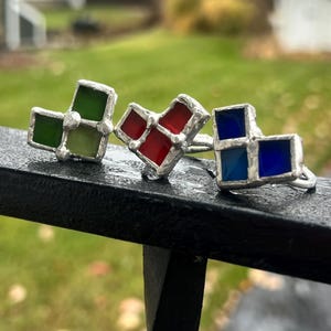 May include: Three handmade rings with square stained glass designs. One ring features green glass, another red, and the third blue. The rings have a silver-colored metal setting and are displayed on a black surface.