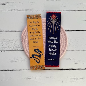 May include: Two bookmarks with inspirational quotes. The first bookmark is yellow with a black dragon design and the quote "We may be small and we may be young but we will shake the world for our beliefs." The second bookmark is blue with a red and gold sun design and the quote "Nothing's worse than a story without an end."