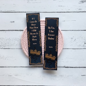 Peut inclure: Deux marque-pages noirs avec des accents dorés et du texte. Le marque-page de gauche dit "Si je ne peux pas être meilleur qu'eux, je deviendrai bien pire." Le marque-page de droite dit "Par toi, je suis à jamais défait." Les deux marque-pages présentent une couronne dorée et un serpent.
