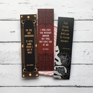 May include: Three rectangular bookmarks with quotes. One is black with gold accents and the text "I'M THE SUN SUMMONER..." The second is burgundy with a pattern and the text "I WILL HAVE YOU WITHOUT ARMOUR..." The third is black with a snake and roses and the text "Not Every Flower Belongs In Every Garden."