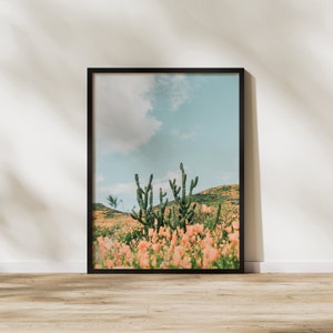 K&ouml;nnte beinhalten: Ein gerahmter Druck eines W&uuml;stenlandschafts mit einer Gruppe von Kakteen im Vordergrund und einem Feld mit rosa Bl&uuml;ten in der Ferne. Der Himmel ist blau mit wei&szlig;en Wolken.