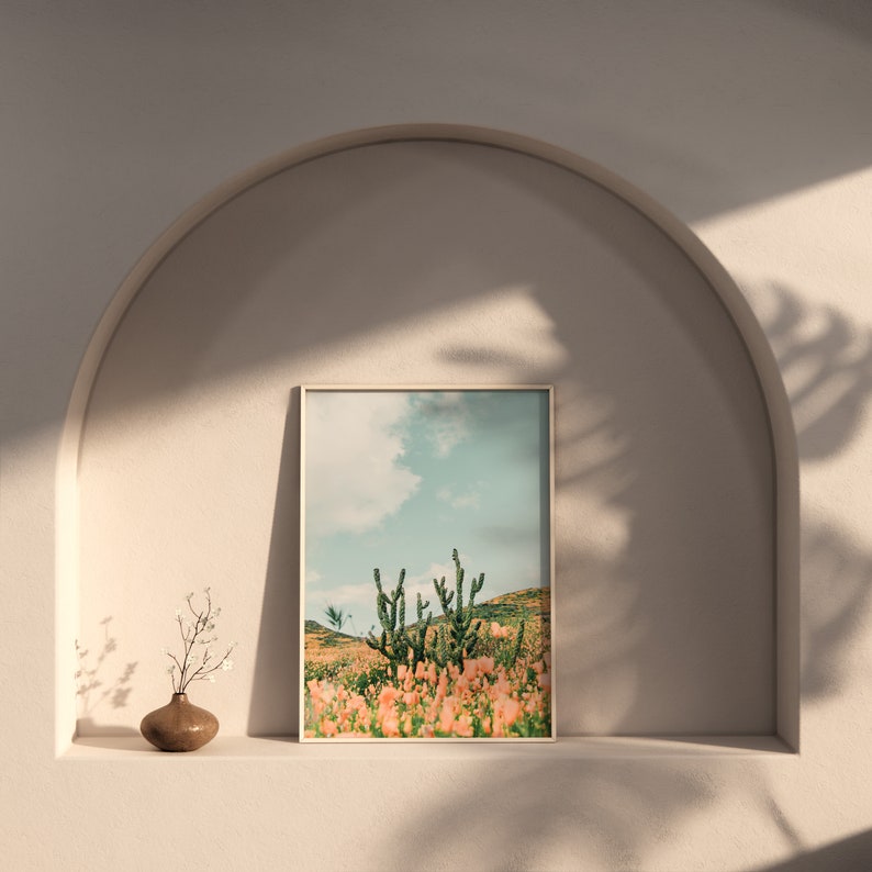 K&ouml;nnte beinhalten: Ein gerahmter Druck eines W&uuml;stenlandschafts mit Kakteen und rosa Bl&uuml;ten. Der Himmel ist blau mit wei&szlig;en Wolken.