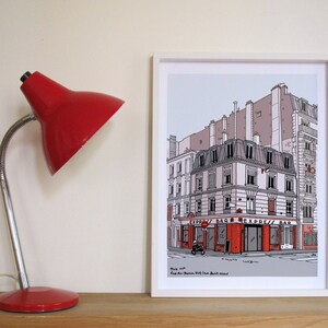 Peut inclure: Une estampe encadrée en blanc d'une illustration dessinée à la main d'une scène de rue parisienne. L'illustration représente un bâtiment avec un auvent rouge et le texte "Express Bar".