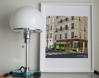 Puede incluir: Una ilustración enmarcada de una escena callejera parisina con un café llamado "Le Troubadour" en la esquina. La ilustración está en un marco blanco y está sobre una mesa de madera con una lámpara blanca.