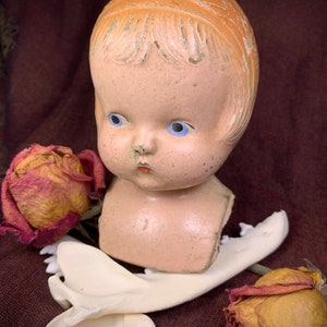May include: A vintage porcelain doll head with blue eyes and a painted smile, resting on a white bone with dried roses on a burgundy fabric background.