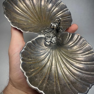 May include: A silver-coloured, two-part dish with a shell-like design. The dish is held open, revealing the interior. A decorative floral element connects the two halves. The dish has a vintage appearance.