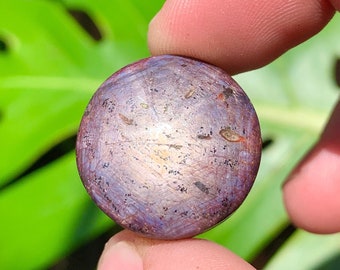 May include: A round, purple and blue gemstone with a star-like pattern and a silver sheen. The gemstone is set in a clear, round cabochon.
