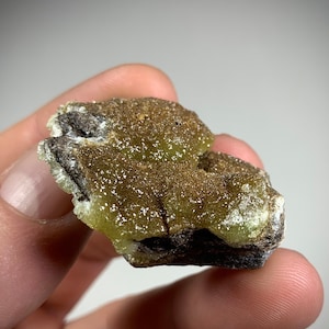 May include: A small, green and brown mineral specimen with a rough, textured surface. The mineral has a crystalline structure and appears to be a type of calcite.