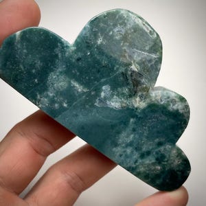 May include: A cloud-shaped stone carving in shades of deep teal and white. The polished surface reveals natural patterns and textures. The object is held in a hand, showcasing the intricate details of the stone.
