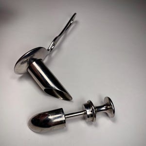 May include: Two stainless steel medical instruments. One is a speculum with a handle and a flared end. The other is a dilator with a rounded tip and a handle. Both instruments are on a white surface.