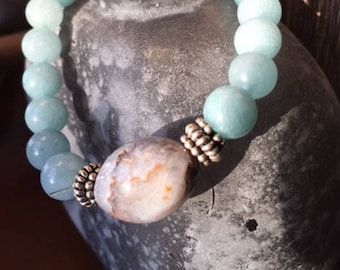 May include: A beaded bracelet featuring light blue and white round beads. The bracelet has a large, oval-shaped, patterned stone in the center, flanked by silver-colored metal accents. The bracelet is displayed on a gray, textured surface.