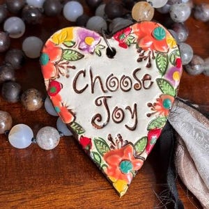 Può includere: Un ciondolo in ceramica a forma di cuore con la scritta "Choose Joy" in lettere marrone scuro, circondato da un bordo floreale dipinto in arancione, giallo, viola e verde. Il ciondolo è infilato su una collana di perline di pietra multicolori.