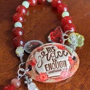 May include: A beaded bracelet featuring red and pale green spherical beads, a central oval ceramic charm with the text "HIS GRACE IS ENOUGH 2 CORINTHIANS 12:9" surrounded by red floral accents, a small silver-toned religious medal, and dangling charms.