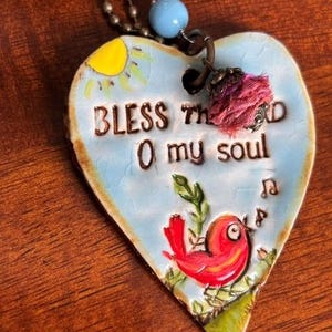 Könnte beinhalten: Ein herzförmiger Keramikanhänger an einer Kugelkette mit hellblauer Craquelé-Glasur, einer gelben Sonne und einem roten Vogel. Der Text lautet BLESS THE LORD O my soul. Eine kleine hellblaue Perle und eine ausgefranste rosa Stoffquaste sind befestigt.