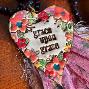 Può includere: Un ciondolo a forma di cuore con la scritta grace upon grace in lettere scure, circondato da fiori dipinti rossi, arancioni e gialli con foglie verdi. Il ciondolo è fissato a una collana di perline nere e a una nappa in tessuto color malva sfilacciato.