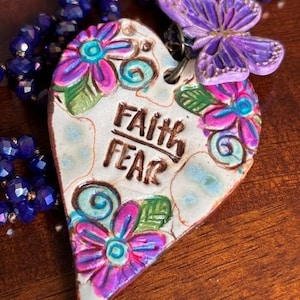 May include: A heart-shaped pendant featuring the text FAITH FEAR in brown lettering, decorated with painted pink and teal flowers and green leaves. A purple butterfly charm is attached to the top, and the pendant hangs from a dark blue beaded necklace.