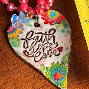 May include: A heart-shaped ceramic pendant featuring the text faith hope and love in brown script, decorated with colourful floral patterns, attached to a red beaded necklace with a small iridescent butterfly charm, next to a yellow ruler.