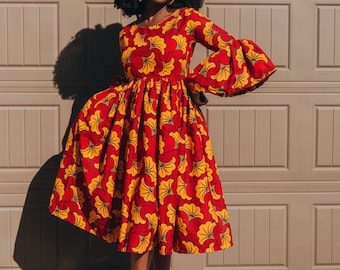 May include: A woman wearing a red and yellow floral print dress with long sleeves and a flared skirt. She is wearing green heeled sandals and has her hair in an afro.