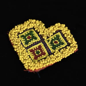 May include: A heart-shaped beaded decoration with a yellow border and a central design of four squares. Each square features a different pattern of green, red, and purple beads. The background is black.