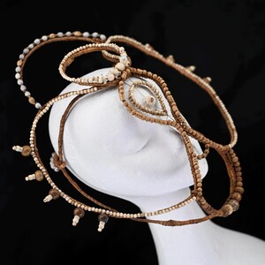 May include: A handcrafted headpiece featuring a complex design of woven brown and tan materials. The headpiece is adorned with small, round, and oblong beads in shades of white, tan, and brown. The design includes multiple loops and strands, creating a layered effect.
