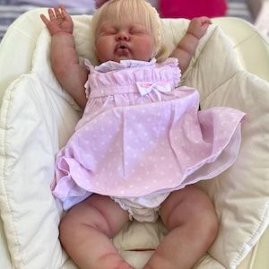 May include: A lifelike baby doll with closed eyes, wearing a light pink dress with white polka dots and a matching bow. The doll has blonde hair and a small purple flower on its head. It is lying in a white cushioned chair.