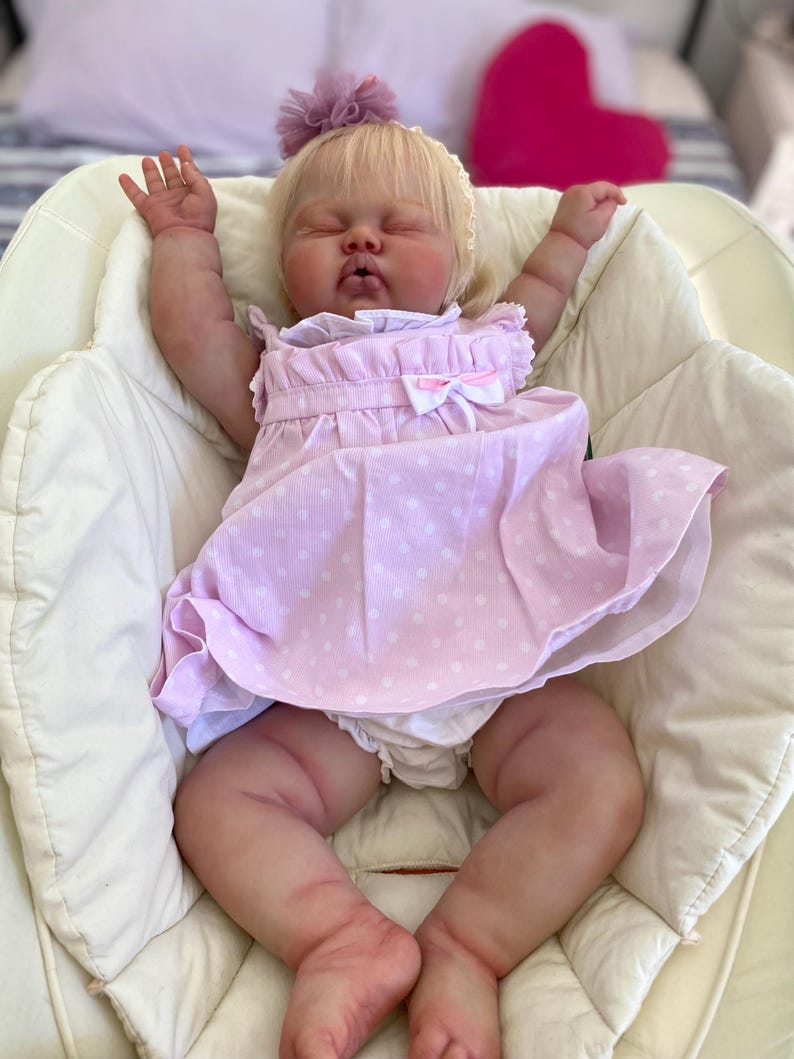 May include: A lifelike baby doll with closed eyes, wearing a light pink dress with white polka dots and a matching bow. The doll has blonde hair and a small purple flower on its head. It is lying in a white cushioned chair.