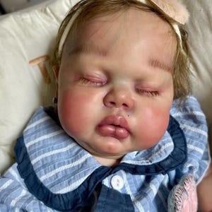 May include: A lifelike baby doll with closed eyes, rosy cheeks, and a small beauty mark on the lip. The doll wears a blue and white striped outfit with a white collar and a headband with a pink bow. The doll is lying on a white pillow.