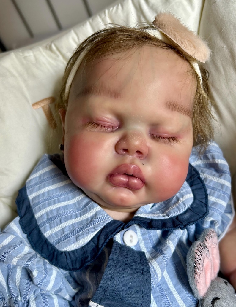 May include: A lifelike baby doll with closed eyes, rosy cheeks, and a small beauty mark on the lip. The doll wears a blue and white striped outfit with a white collar and a headband with a pink bow. The doll is lying on a white pillow.
