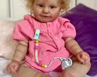 Puede incluir: Una muñeca bebé realista con cabello rubio rizado y ojos marrones, vestida con un body rosa. La muñeca tiene un chupete sujeto a una cadena de cuentas. La muñeca está sentada sobre una sábana blanca con un cojín morado en el fondo.