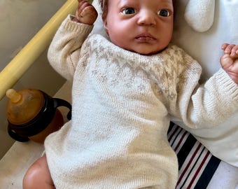 Puede incluir: Una muñeca realista de piel oscura y cabello castaño rizado, que lleva un pelele de punto color crema con un sutil patrón de estrellas en el pecho y una pequeña diadema de flores. La muñeca está colocada cerca de un biberón marrón.